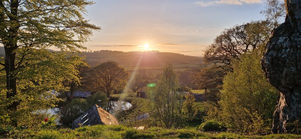 A bright yellow sun setting over a valley with shards of white light hitting a slate roof in the foreground. The trees and grass all bathed in a soft yellow.